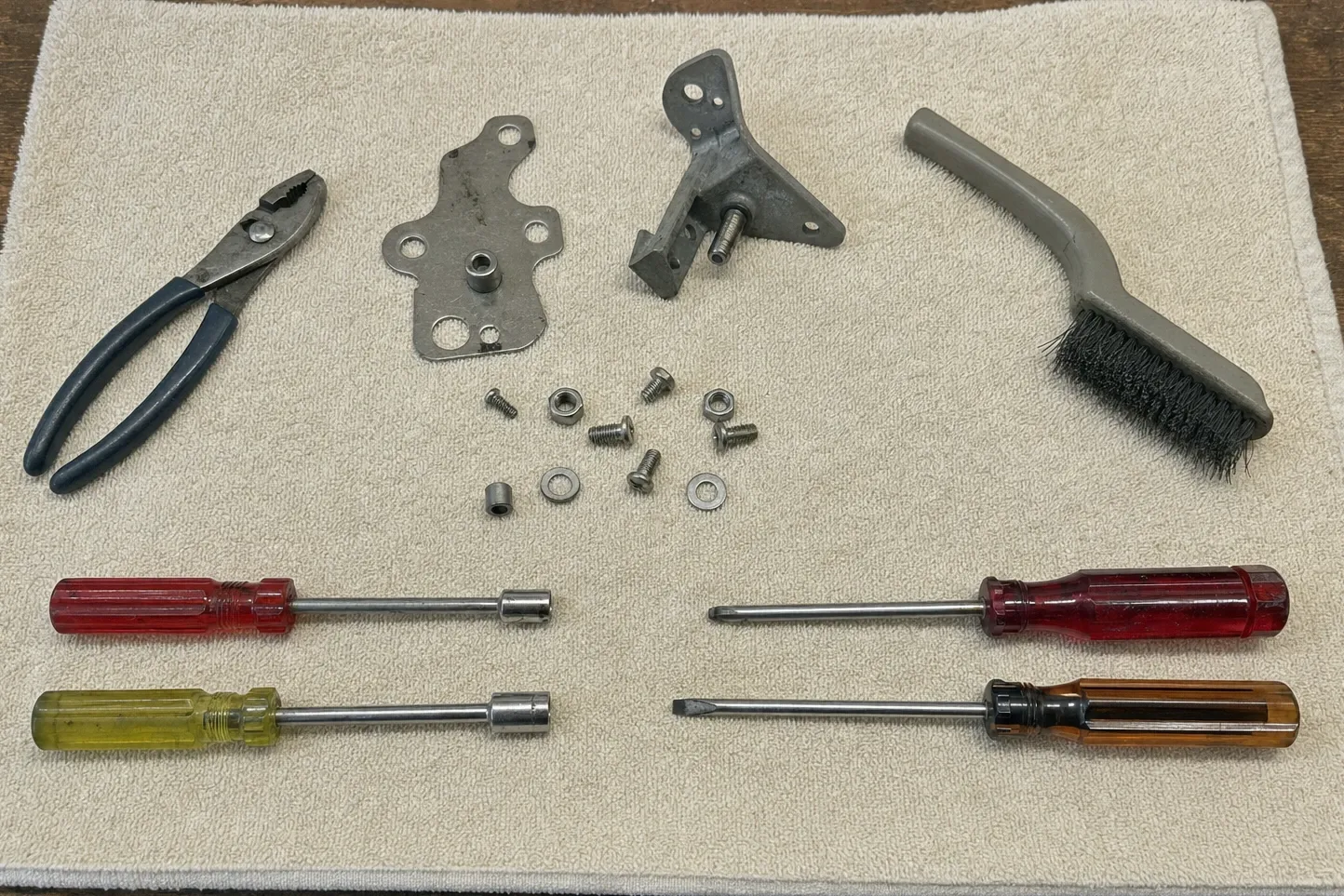 Slip-joint pliers, four drivers and a wire brush laid out with two flat link plates, an angle bracket and loose fixings
