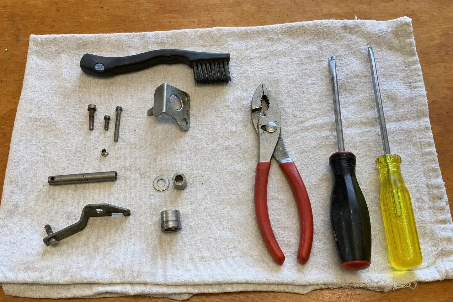 Slip-joint pliers, screwdrivers and a wire brush on cloth beside a cast lever arm, a roll pin and a threaded collar